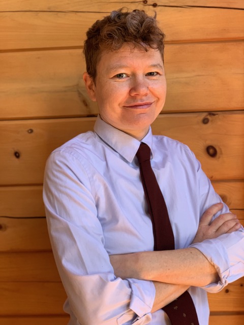 Hartman has short hair and a smile, arms crossed and wears a brown tie and a grey shirt