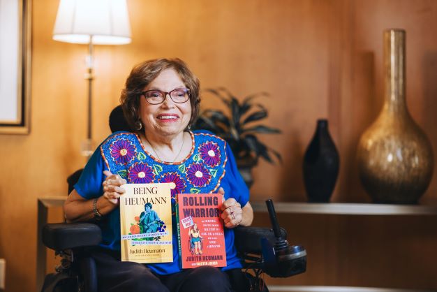 A photo of Judy Heumann holding her two books, Being Heumann and Rolling Warrior. Judy is a white woman with short brown hair who uses a wheelchair. She is wearing glasses, a bright blue shirt with pink embroidered flowers, and black pants. The book covers are yellow and red.