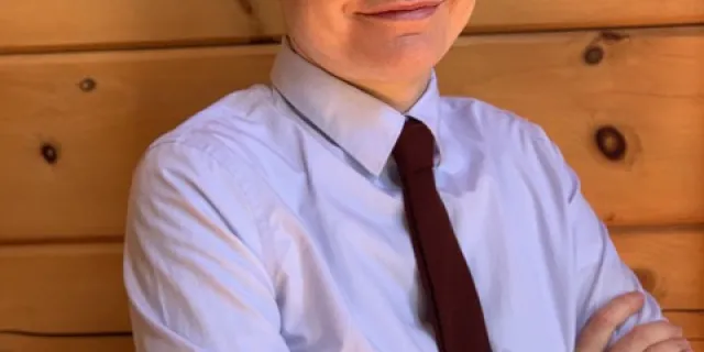 Hartman has short hair and a smile, arms crossed and wears a brown tie and a grey shirt